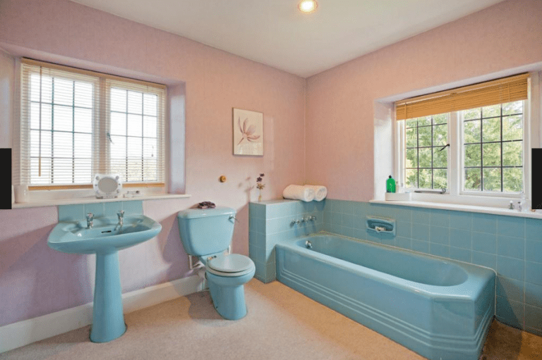 A vintage-style bathroom with pink walls and light blue fixtures, including a pedestal sink, toilet, and bathtub. Two large windows with blinds provide natural light, and a small floral artwork hangs above the toilet.